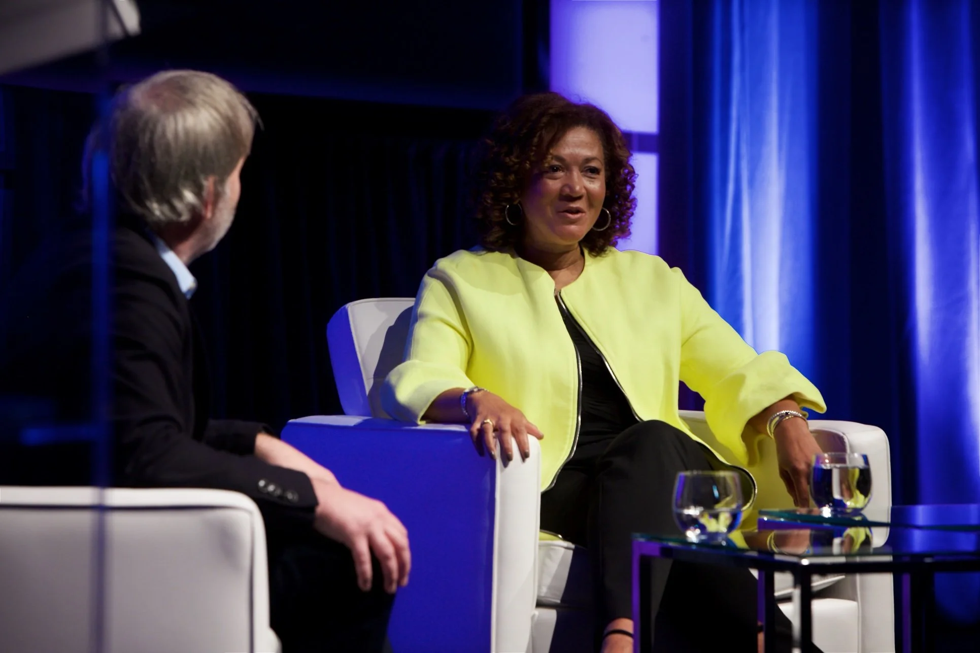 A woman in a bright yellow jacket and black outfit sitting and speaking during an interview or panel discussion, with a man in a dark suit listening.