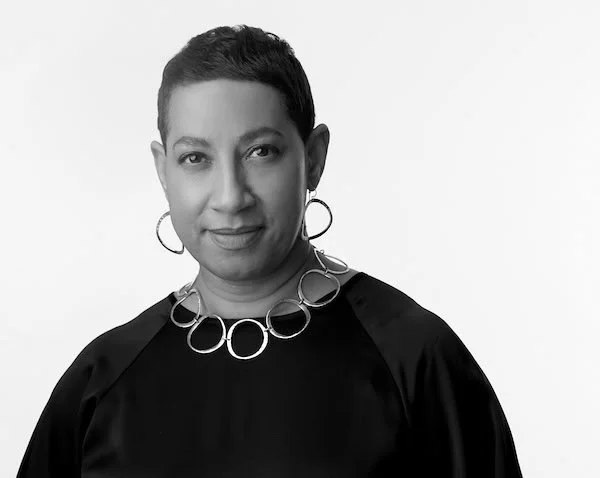 Black and white portrait of a woman smiling, short dark hair, wearing a textured jacket and hoop earrings against a dark background.