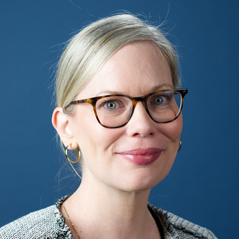 A woman with blonde hair, glasses, and earrings smiling at the camera, with a plain light-colored background.