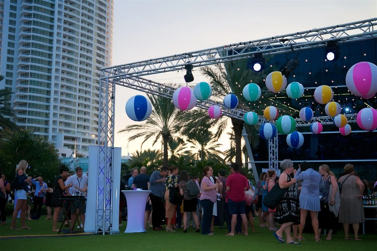 People gathering outdoors at a social event with a stage decorated with colorful beach ball-shaped lanterns, high-rise buildings, and palm trees in the background during sunset.