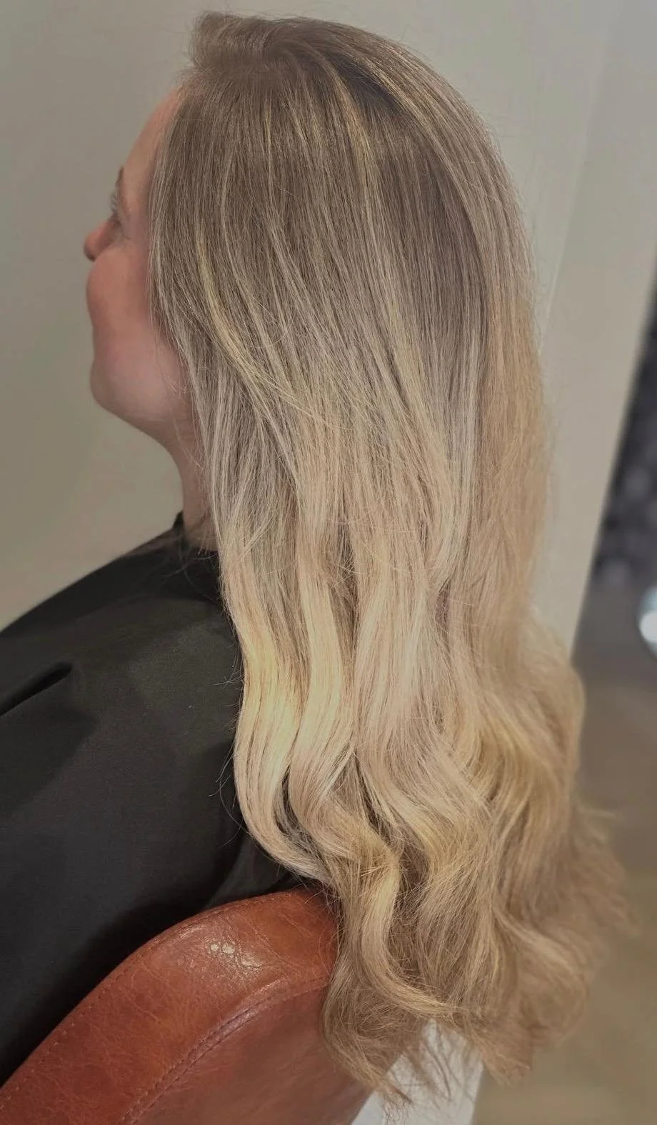 Side view of a woman with long, wavy, blonde hair, seated on a brown leather chair.
