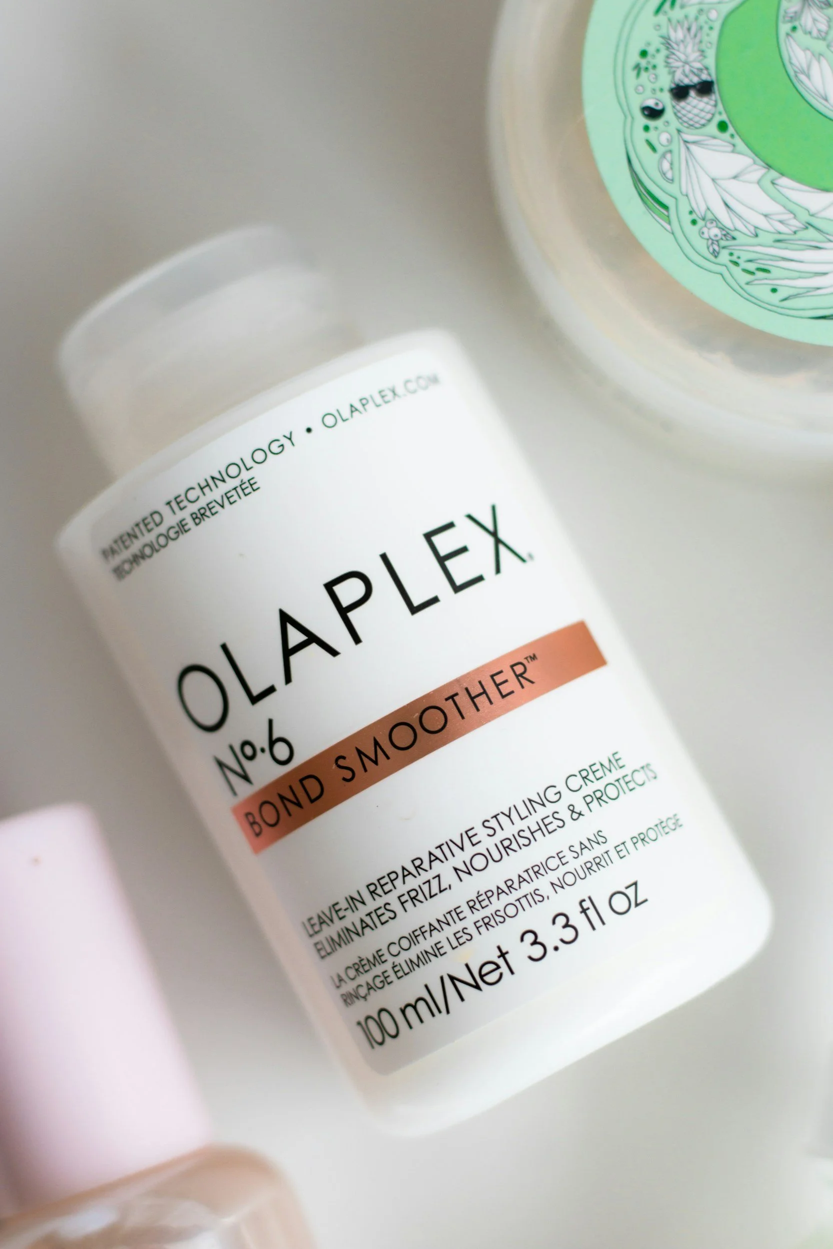 Close-up of a white bottle of Olaplex Bond Smoother hair treatment with black text and a copper-colored stripe, alongside a partially visible container with a green label in the background.