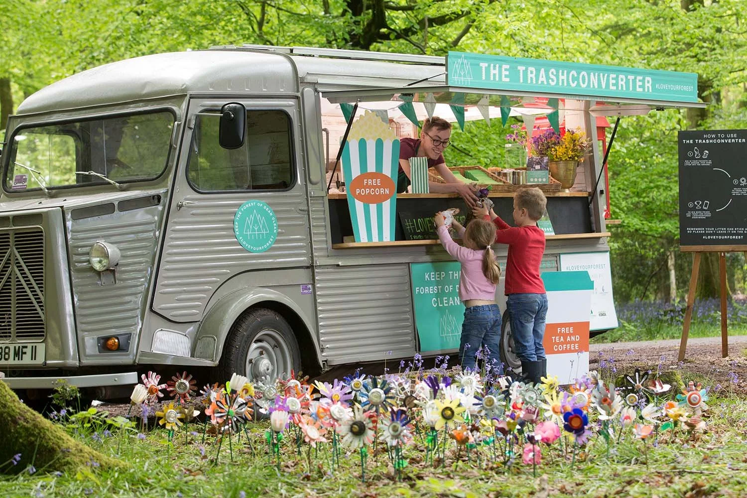 The trashconverter is parked in a forest. A man is swapping two children's trash that they have collected for treats.