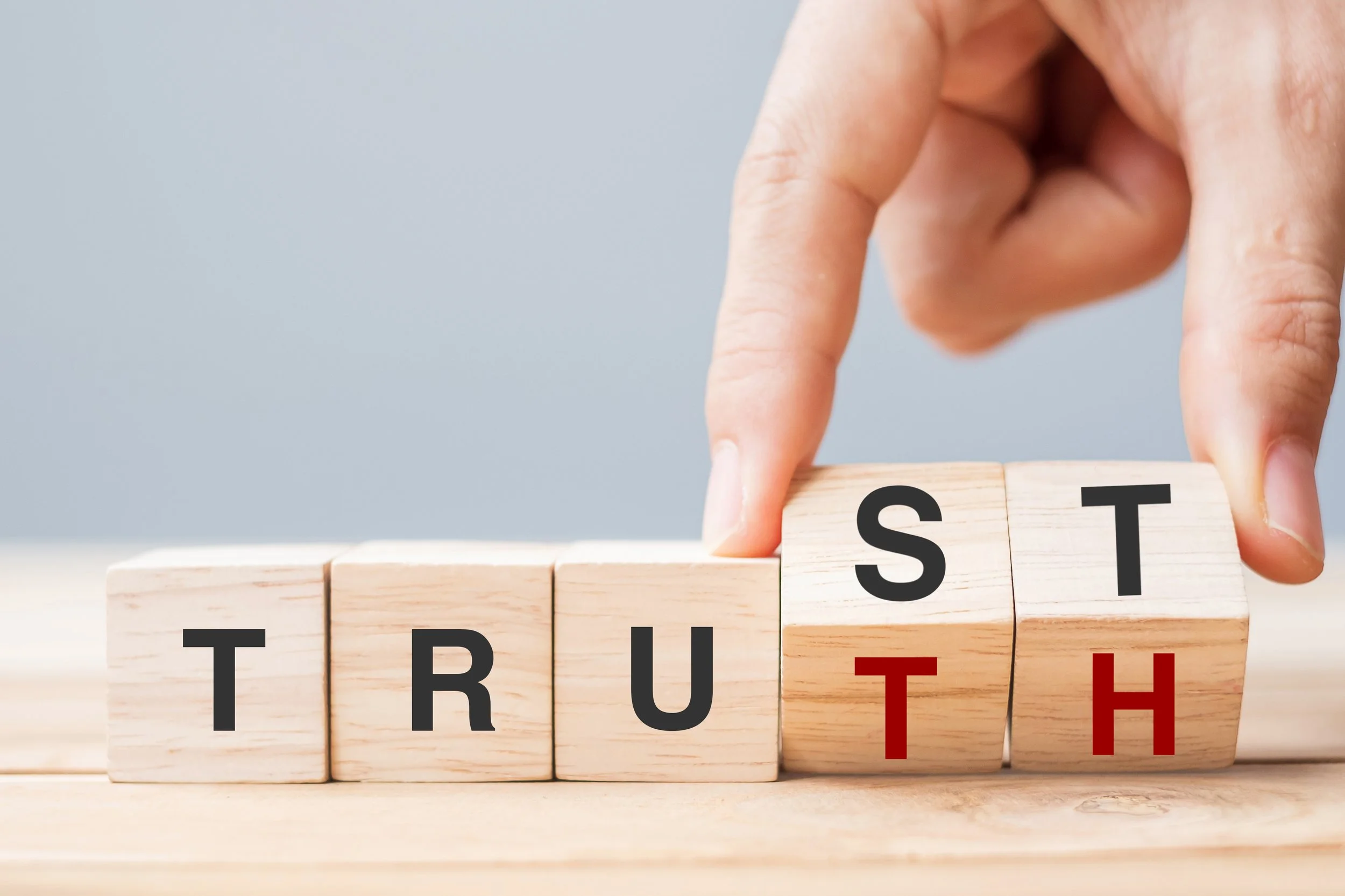 A series of blocks spell out the word trust in black letters, A hand is turning the last two blocks to reveal red letters T and H, changing the word from Trust to Truth.