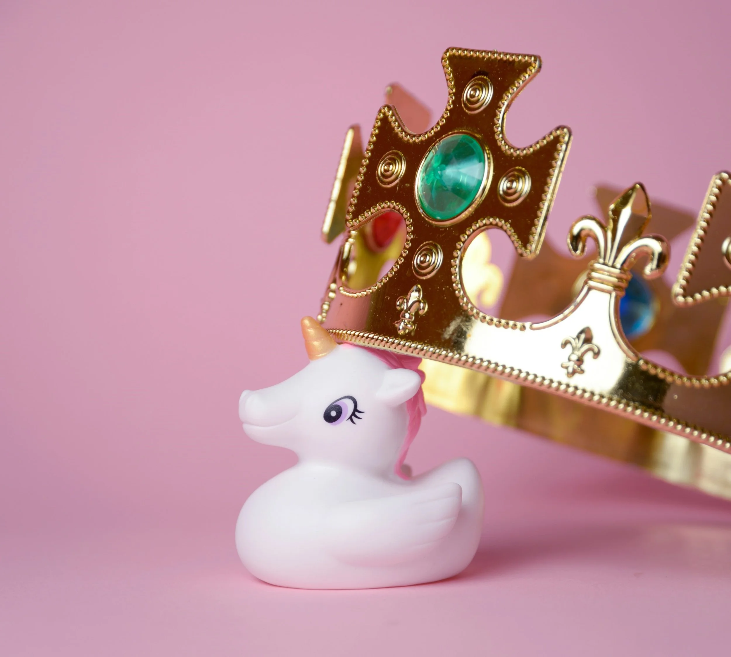 A small rubber unicorn (like a rubber duck but  white and with a horn and pink mane) is sat centre of the picture against a pink background. A giant plastic croen with emaralds and rubies on it is perched on her head. It is twenty times the size of t