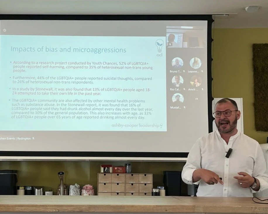 Matt is stood in front of a screen that shows the impacts of bias and microaggressions. He's wearing a white shirt, glasses and a mic and is stood behind a counter top.