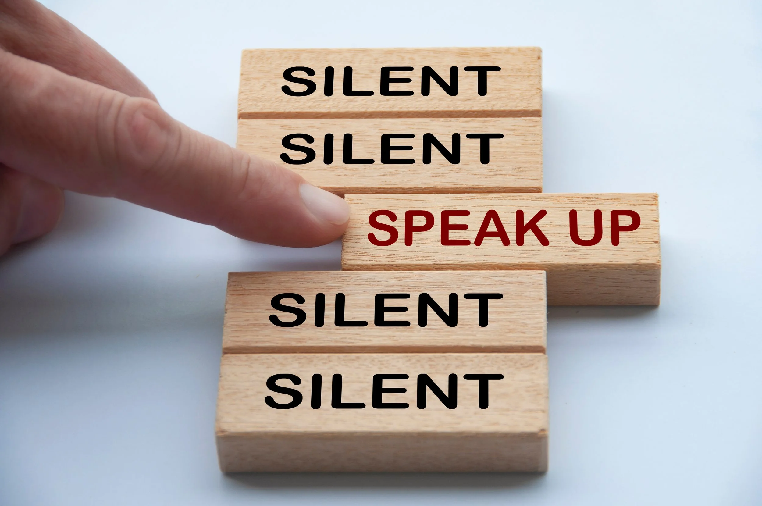 Five blocks are stacked, the top and bottom two say Silent in black letters, the middle one says Speak Up in red letters - this one is being pushed out by a finger from the left hand side..