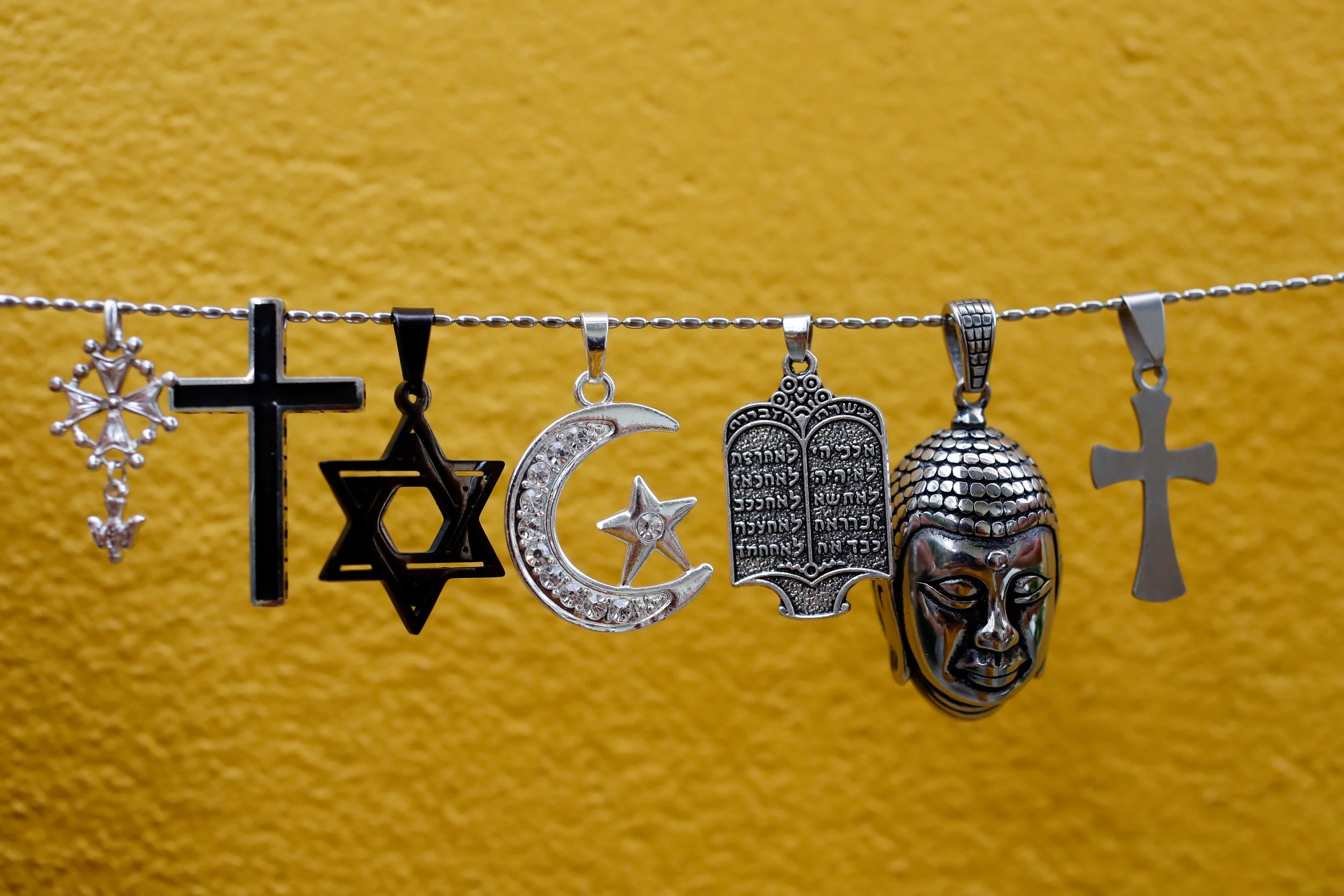 A gold background, in the foreground a chain hangs with a number of religious pendants hanging from it, including crosses, a star of David, a Buddhist figure, a Torah, and a Crescent moon and star.