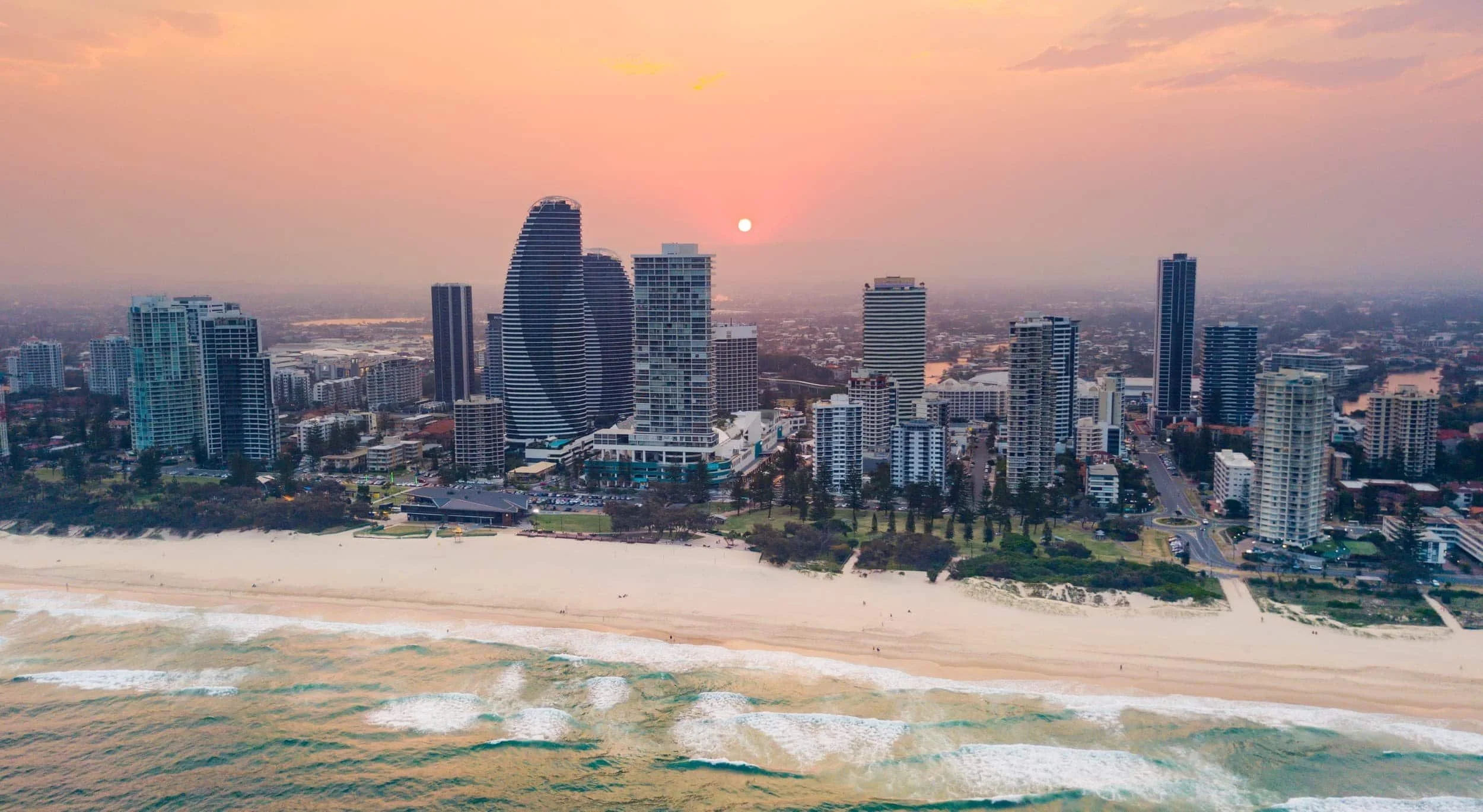 A city skyline at sunset with high-rise buildings along a sandy beach, ocean waves in the foreground, and a partly cloudy sky with the sun near the horizon.
