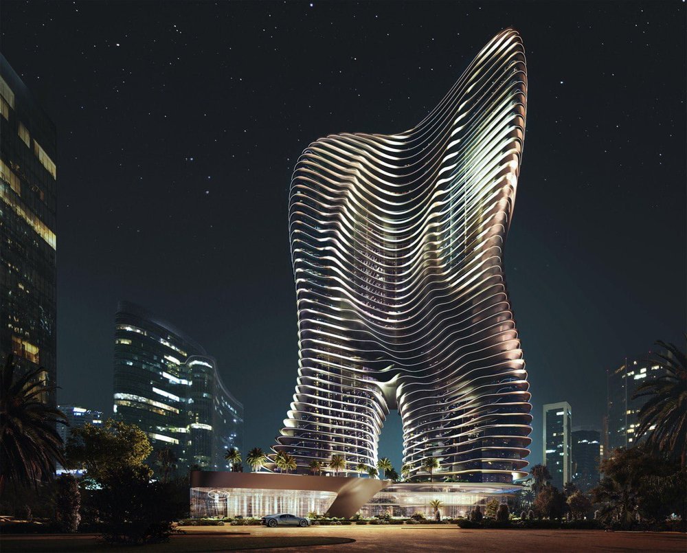 Night view of a futuristic high-rise building with curving, layered design, illuminated by exterior lighting, with surrounding modern skyscrapers and palm trees.