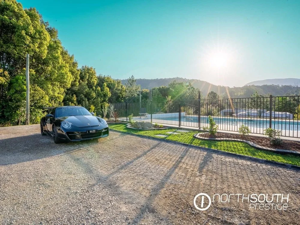 A black Porsche sports car parked on a gravel driveway near a fenced swimming pool with a landscape of trees and hills under a clear sky with the sun shining.