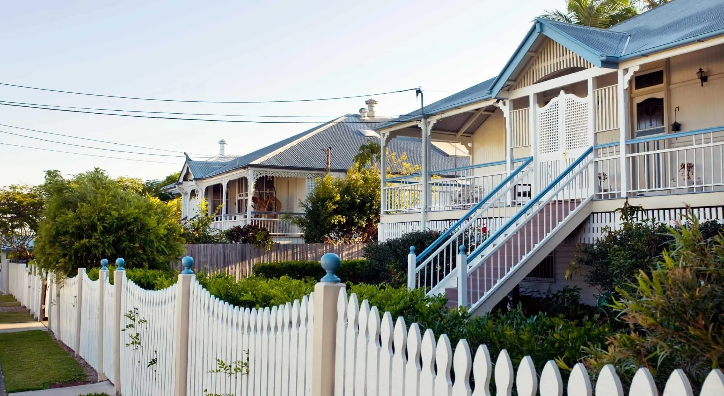 A row of Victorian-style houses with elaborate trim and porches, behind a white picket fence, with gardens and trees in the foreground, under a clear sky.