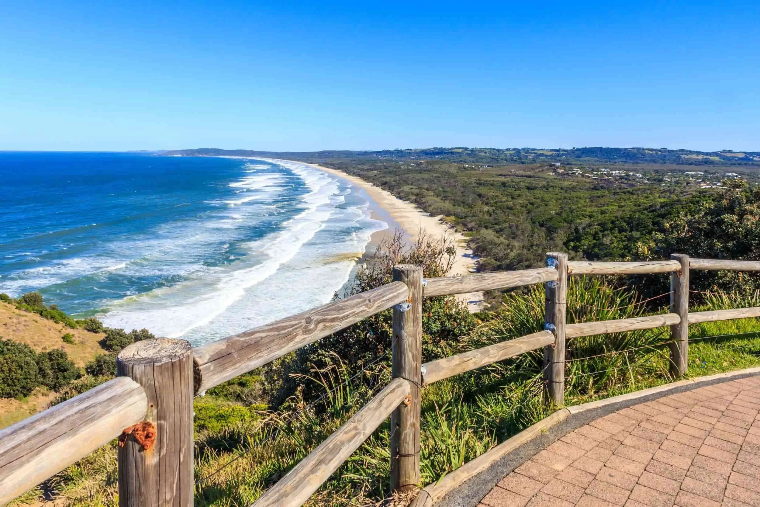 View of a coastline with sandy beach, blue ocean waves, green hills, and a wooden fence in the foreground under a clear blue sky.