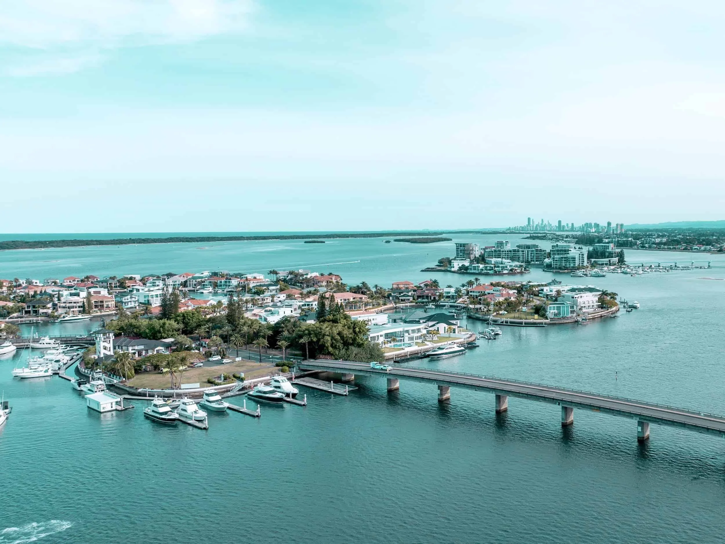 Aerial view of a coastal cityscape with water, boats, residential buildings, and a distant skyline under a cloudy sky.