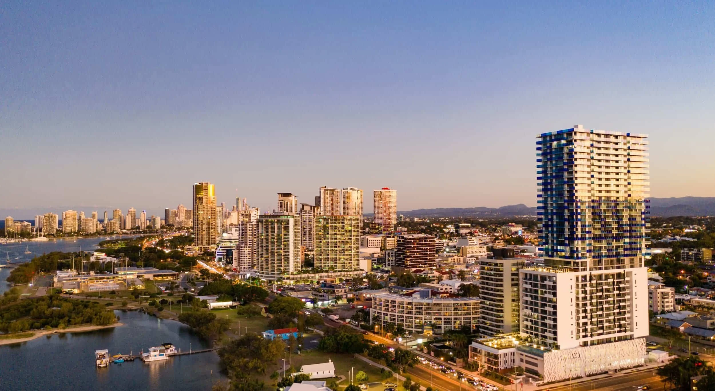 A city skyline at sunset with high-rise buildings, a river with boats, and a mountain range in the background.