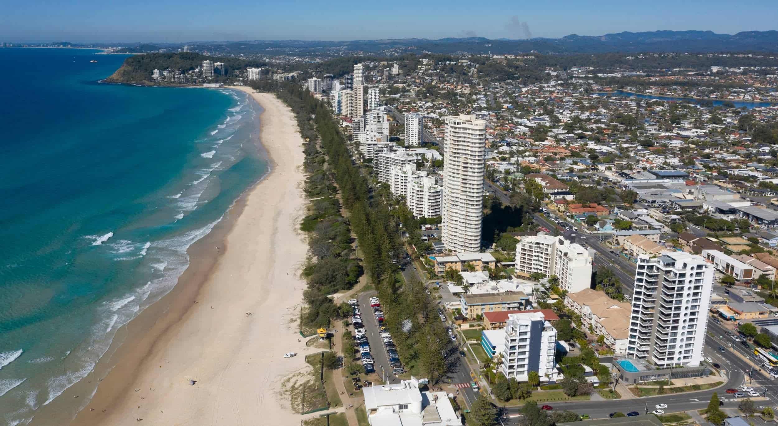 Aerial view of a coastal city featuring tall buildings along a sandy beach with turquoise water, and hills in the background.