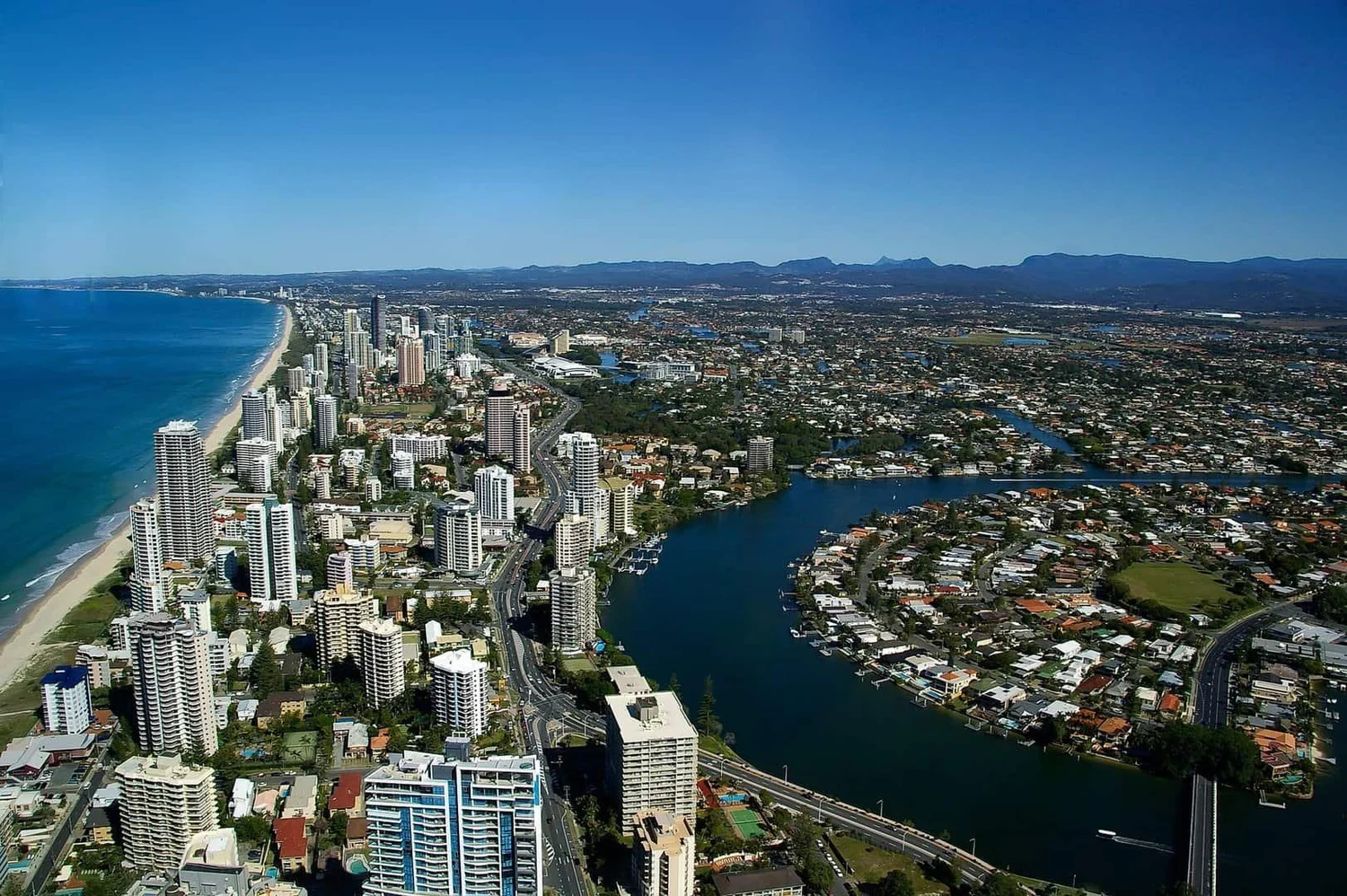 Aerial view of a coastal city with high-rise buildings along the beach, a river running through the city, and mountains in the background on a clear, sunny day.