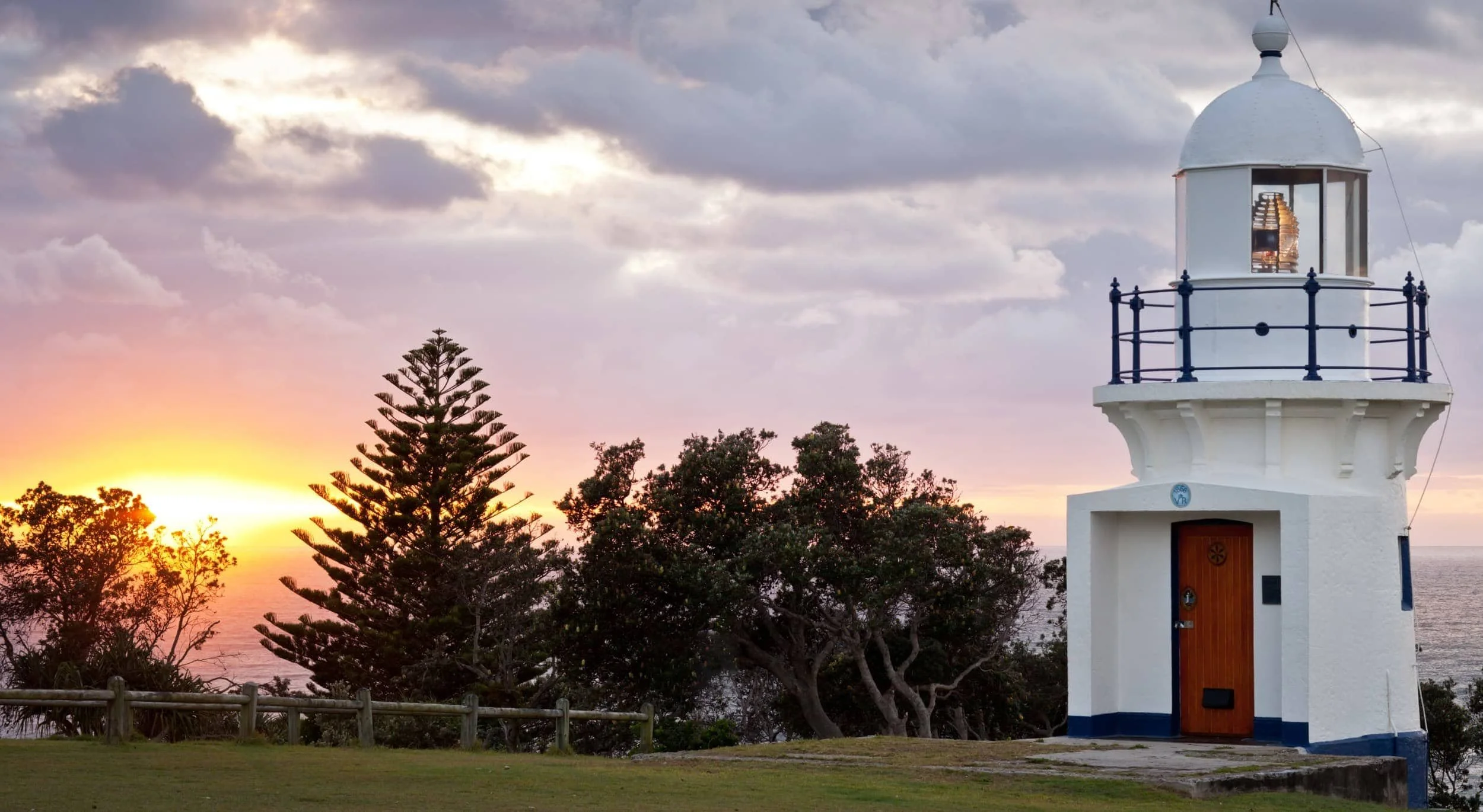A white lighthouse with a black railing around its top, located near the ocean during sunset, with trees and a fence in the foreground and a cloudy sky overhead.
