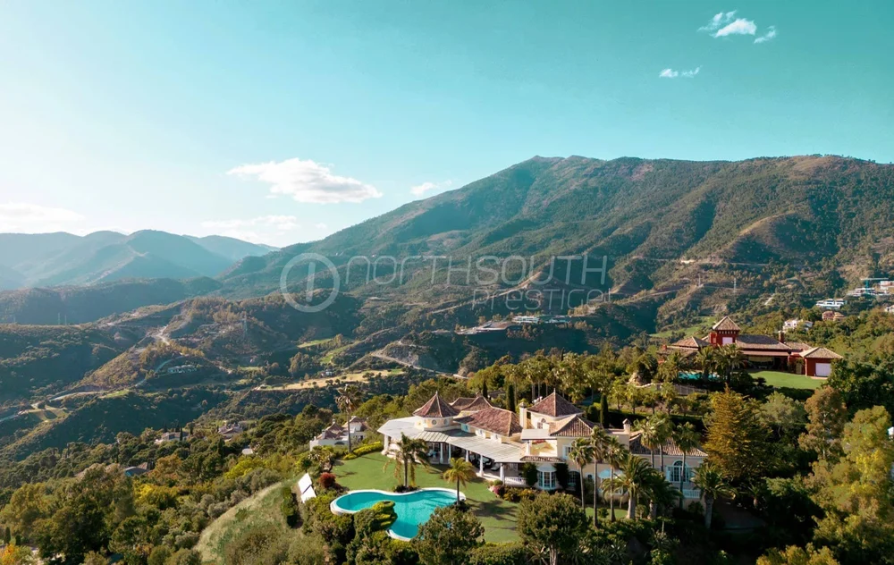 Luxury house with a swimming pool in a lush green valley surrounded by mountains under a bright blue sky with some clouds.