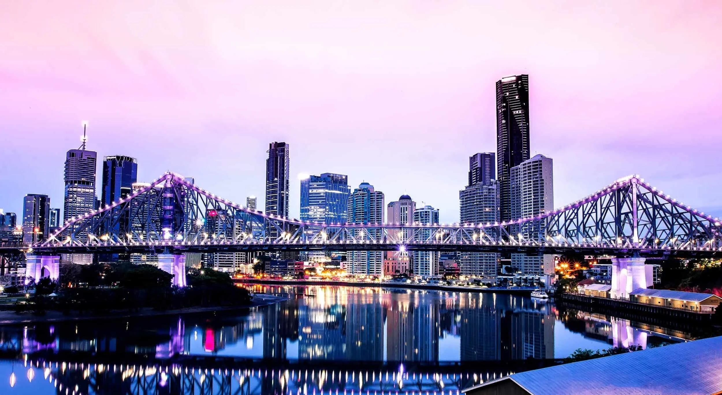Nighttime city skyline with illuminated skyscrapers and a bridge over a river, reflected in the water.