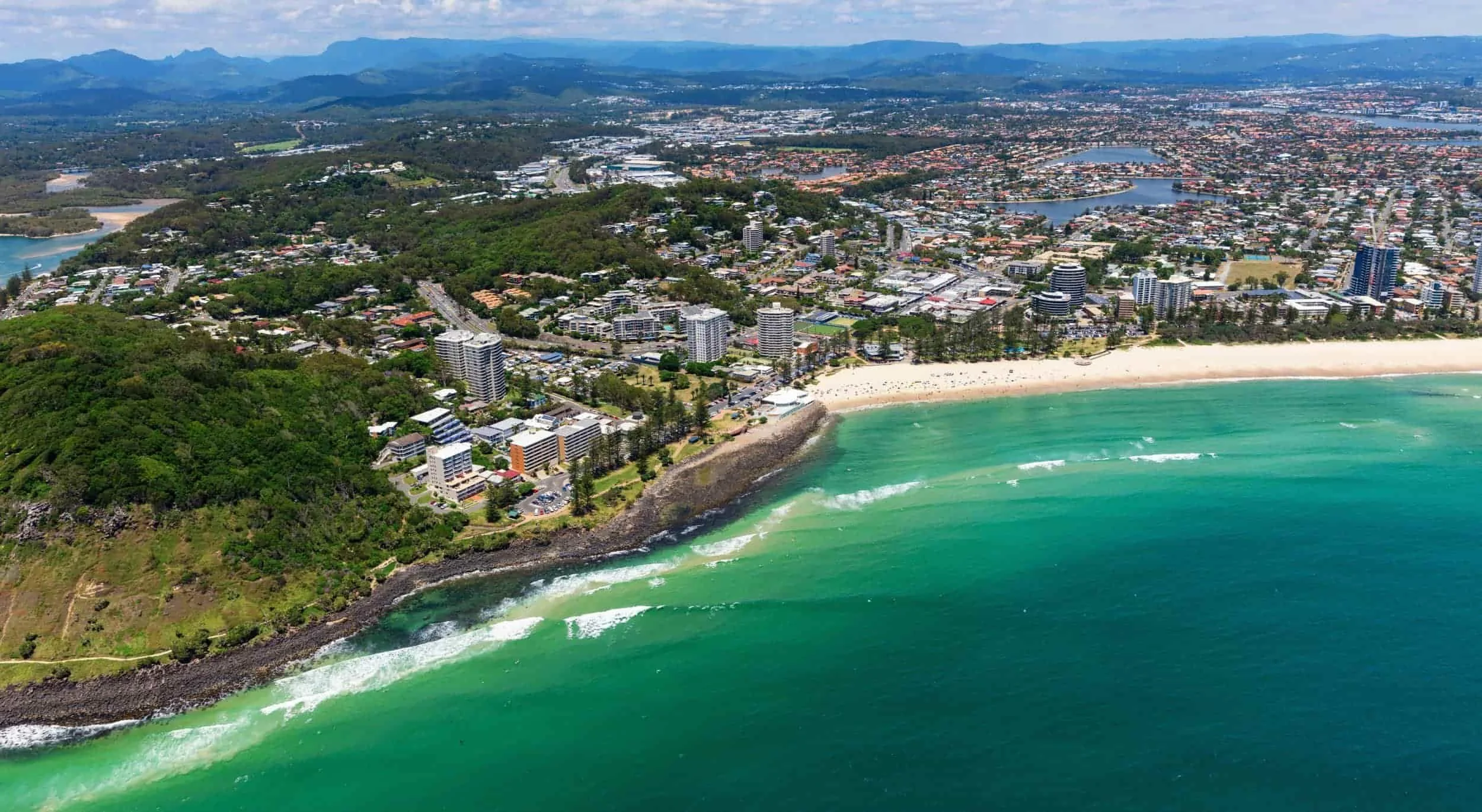 Aerial view of a coastal city with high-rise buildings, a sandy beach, and turquoise ocean waves. Green hills and city streets are visible, with lakes and mountains in the background.