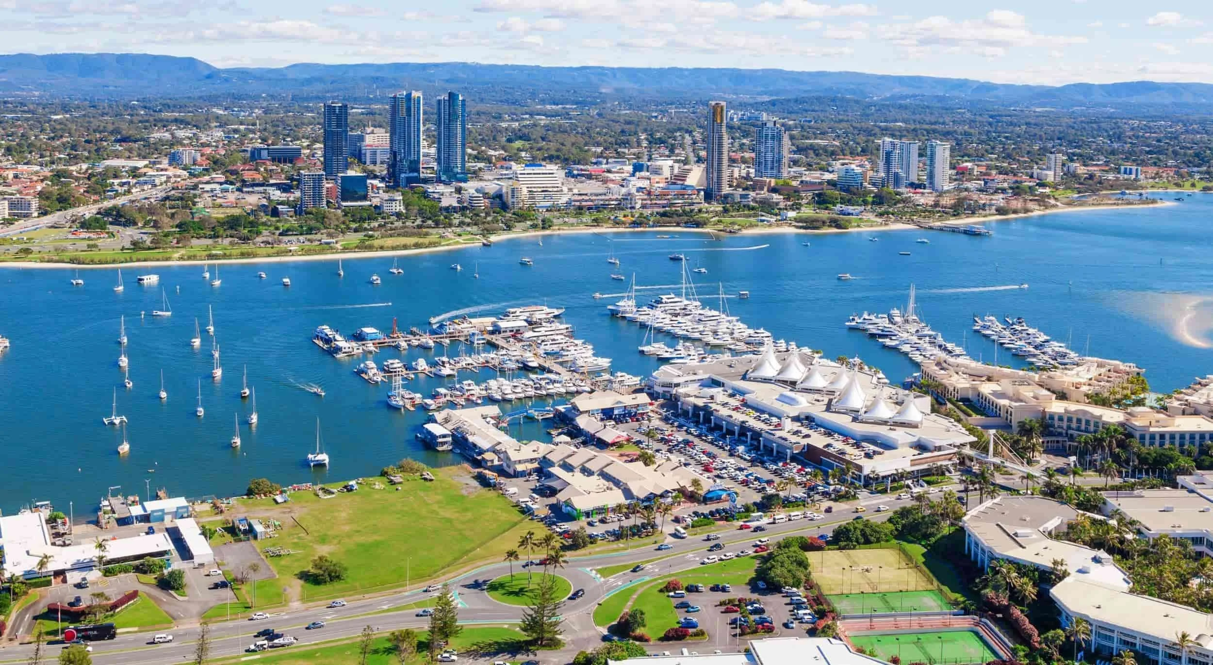 Aerial view of a city skyline with tall modern buildings, a bay with numerous sailboats and yachts, and a waterfront area with white tents and a marina. There are green parks, roads, and residential areas surrounding the water.