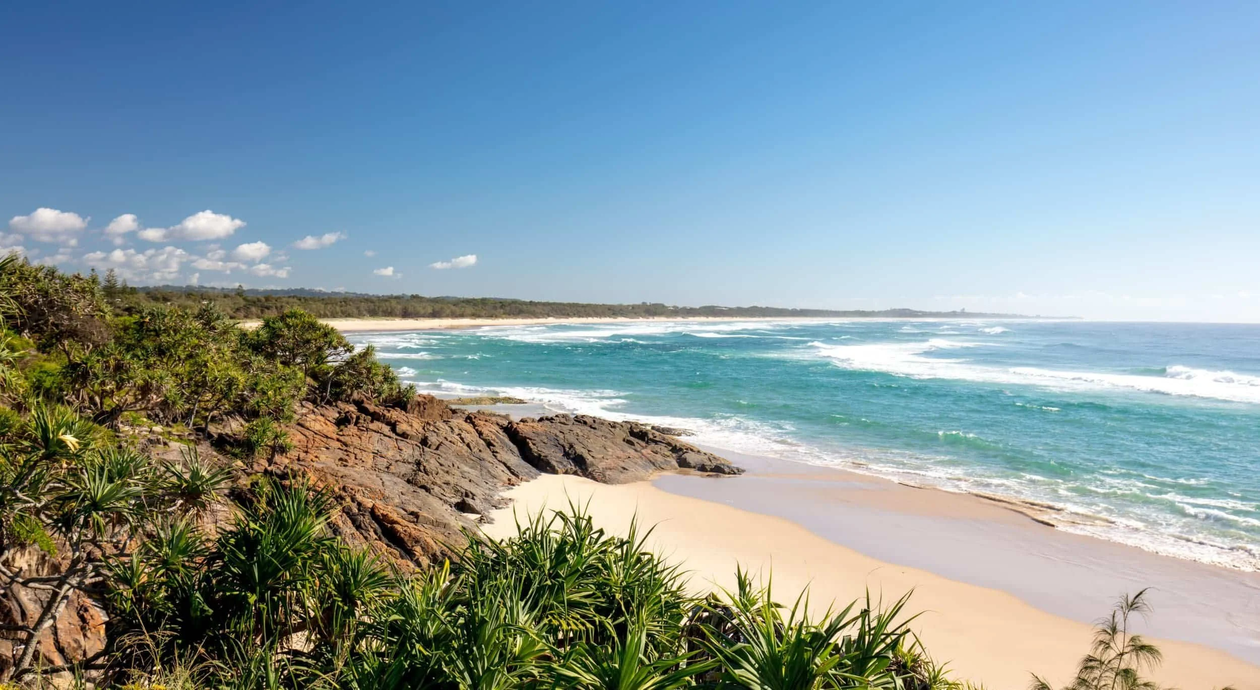 A scenic view of a beach with white sand, rocky cliffs covered with green plants, and the ocean with waves under a clear blue sky with some fluffy clouds.