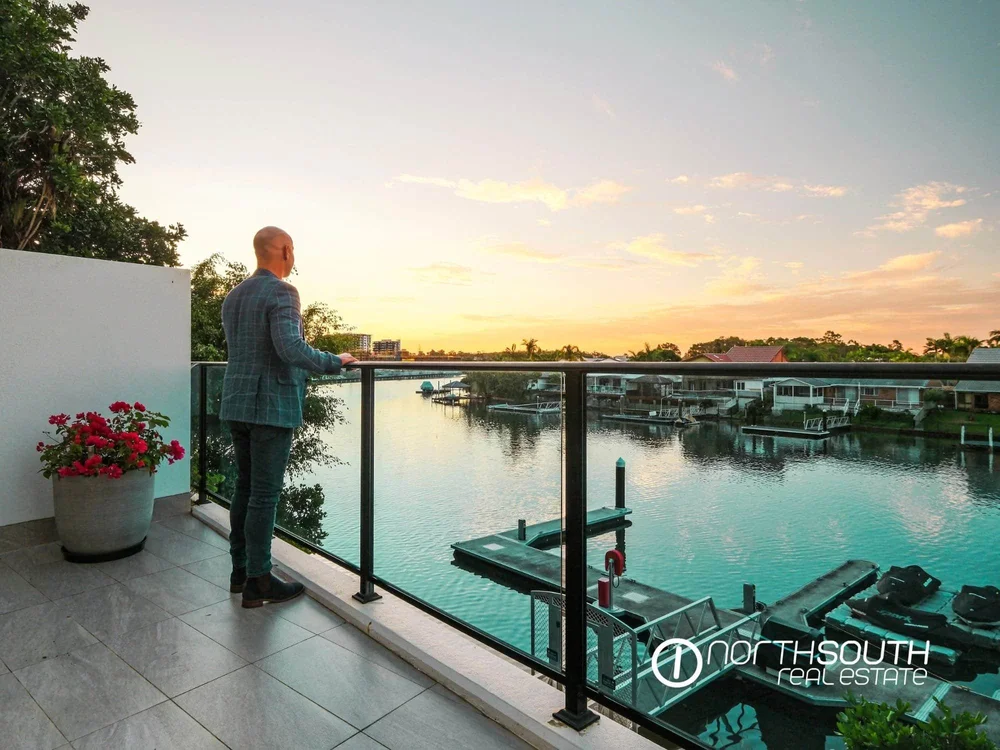 Overlooking the water canal during sunset from a property balcony. The balcony has a glass railing, and there are some boats on the water. There are trees and buildings in the background.