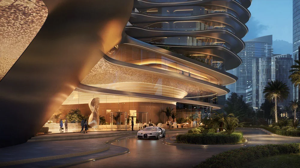 Nighttime view of a modern skyscraper with curved architectural design, glass windows, and surrounding city buildings. A luxury sports car is parked outside, and there are people walking near the entrance. The area is landscaped with palm trees and d