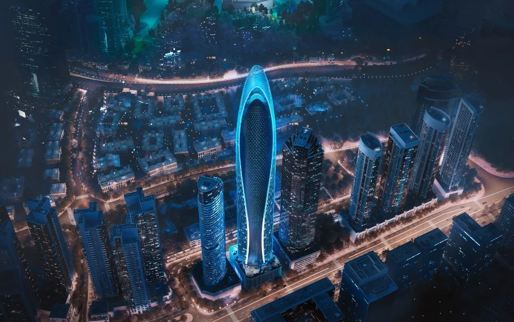 A nighttime aerial view of a city with tall skyscrapers, including a distinctive oval-shaped illuminated building in the center, surrounded by roads and other buildings.
