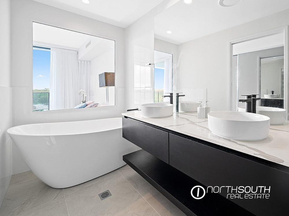 Modern bathroom with a white bathtub, large mirror, double vessel sinks, black faucets, black cabinetry, and a large window with a view of the outdoors.