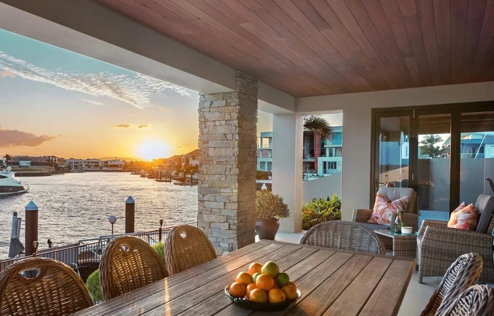 A covered outdoor patio area overlooking the water at sunset. The patio has wicker chairs with cushions, a wooden table with a bowl of fruit, potted plants, and sliding glass doors leading inside.