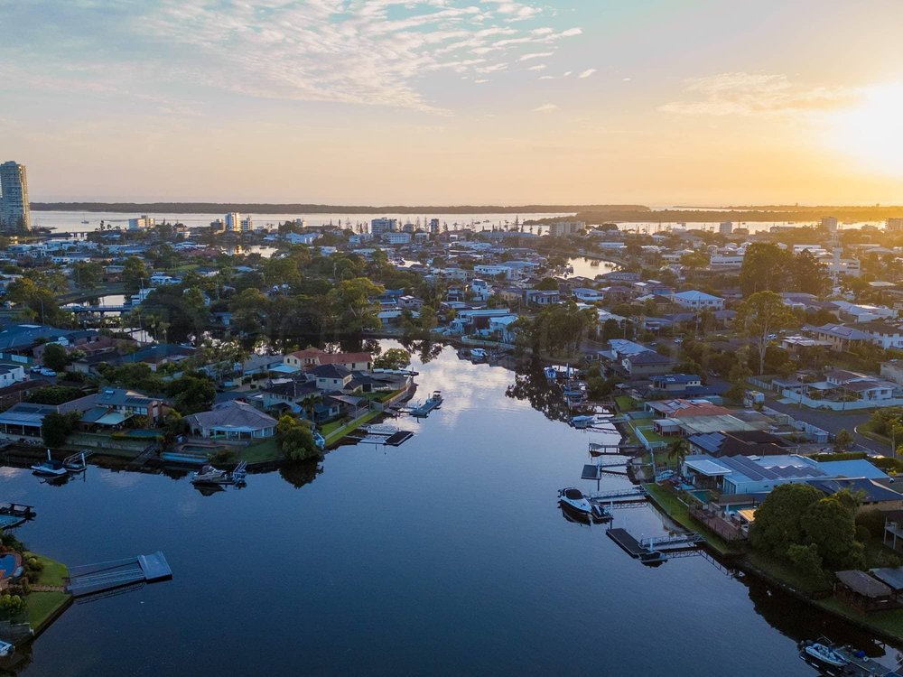 Sunset over a suburban neighborhood with houses, canals, and boats, near a body of water with a large river or lake in the background.