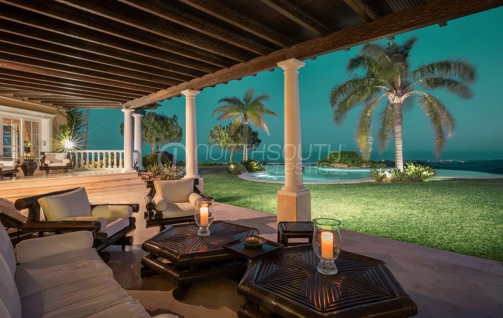 The villa lit from within on a warm evening, seen across its terraced gardens.