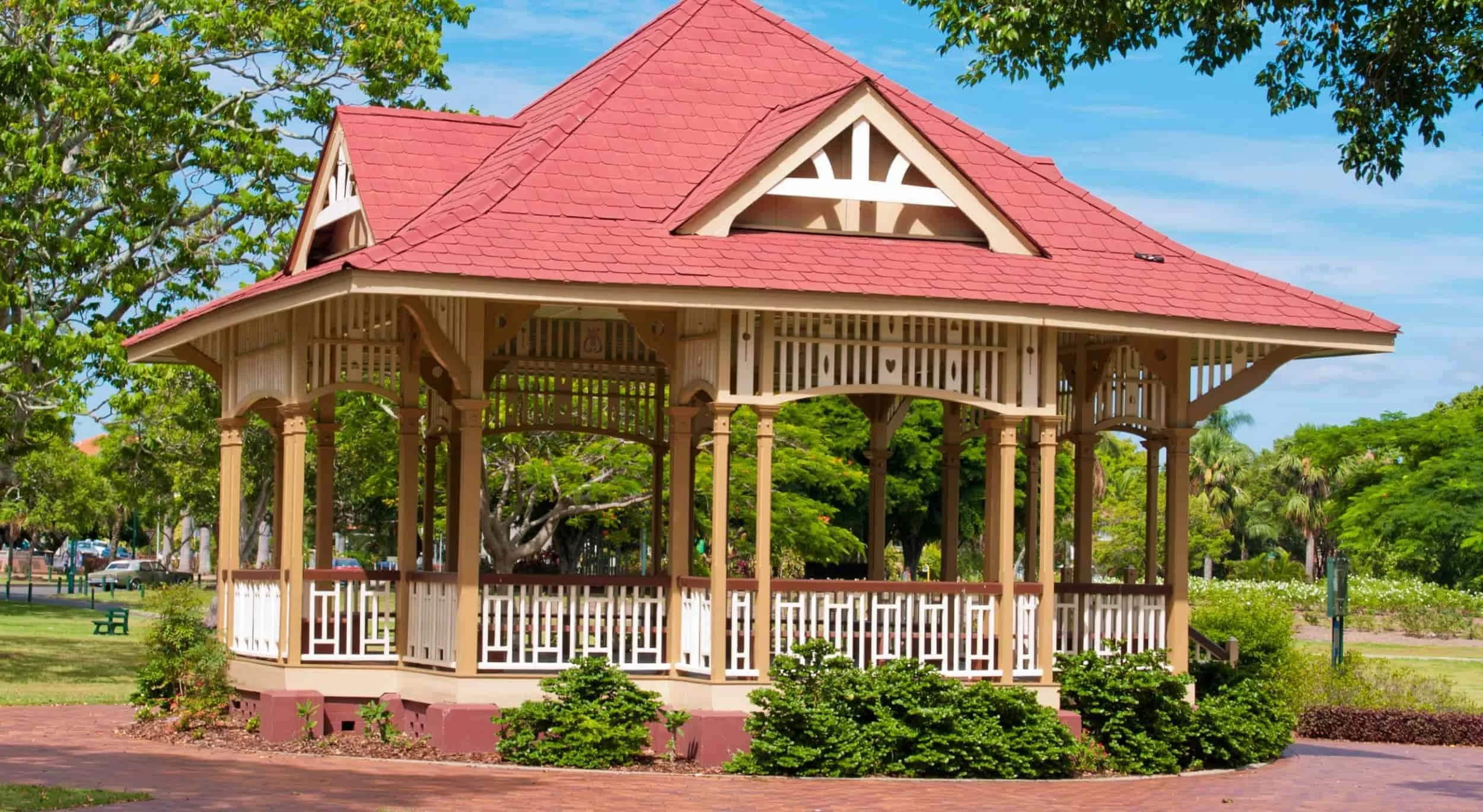 A decorative wooden gazebo with a red shingled roof, located in a park with green trees, grass, and bright blue sky.