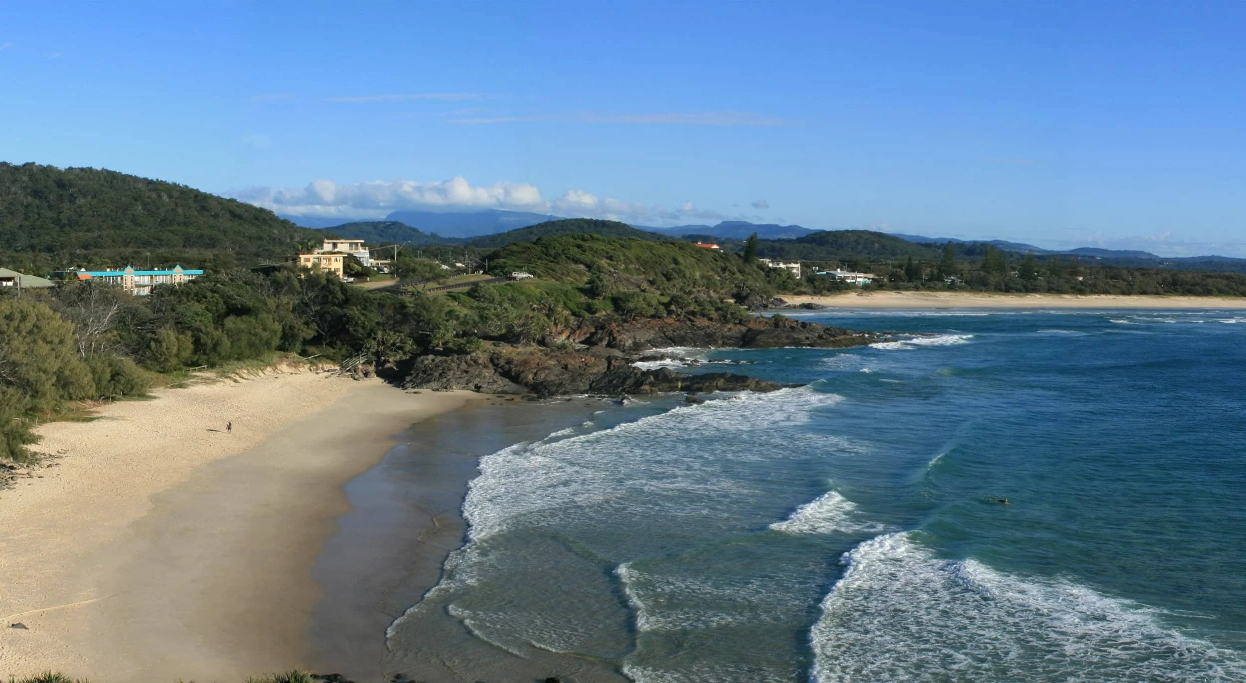 A scenic coastal view with a sandy beach, ocean waves, green hills in the background, and a few buildings on the hillside under a clear blue sky.
