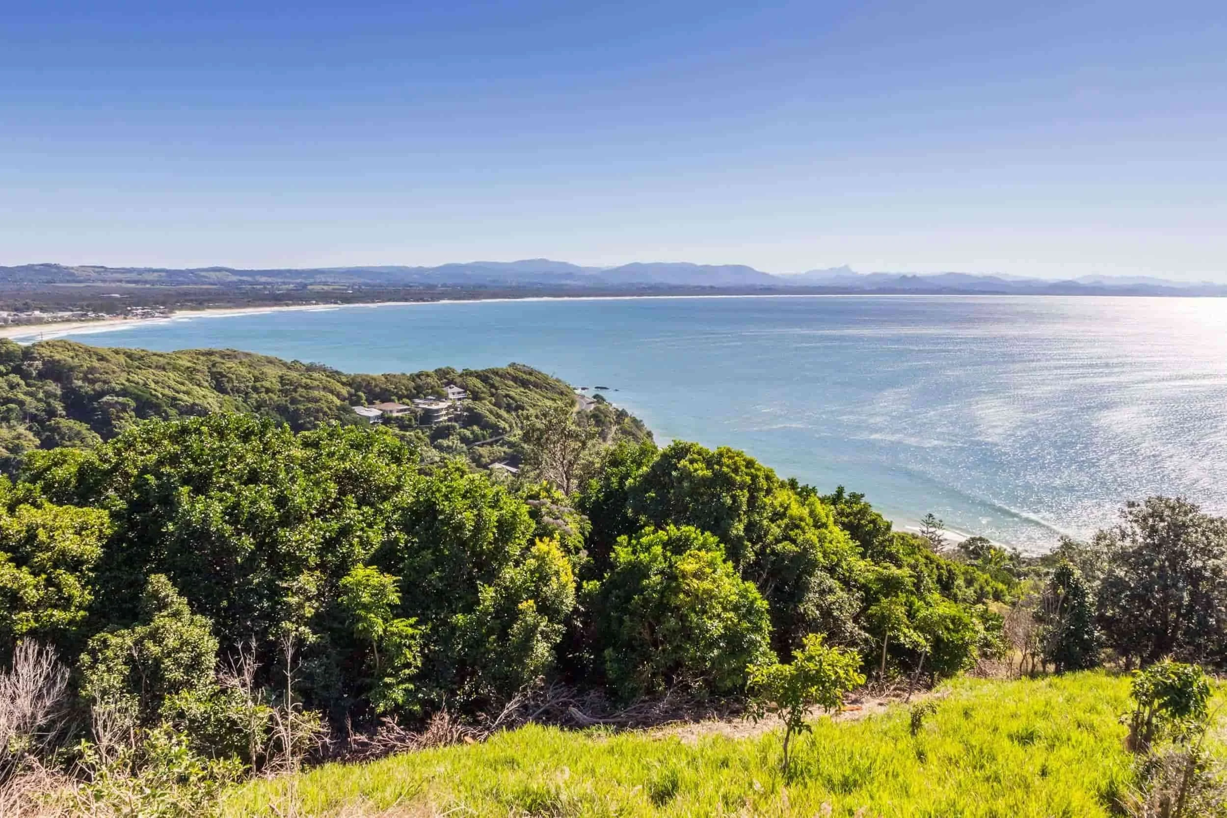A scenic view of a coastal landscape with lush green trees and vegetation in the foreground, a calm blue ocean, and distant mountains under a clear blue sky.