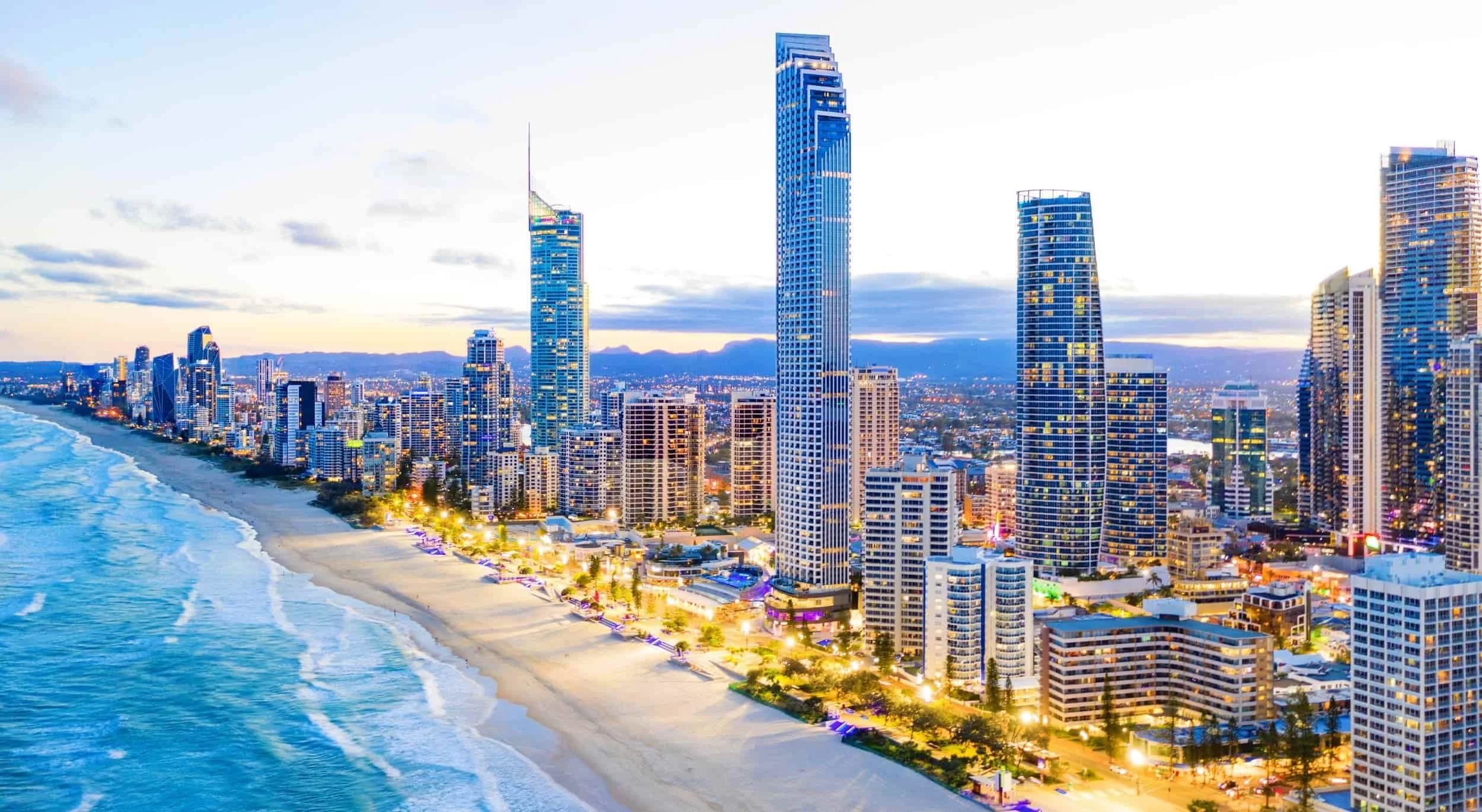 Aerial view of a city skyline with tall modern buildings along a beach at sunset, with ocean waves on the left and illuminated streets.