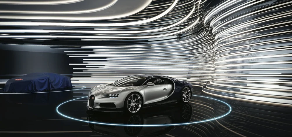 A silver luxury sports car displayed in a modern futuristic showroom with curved, illuminated metallic walls and a circular light strip on the floor.
