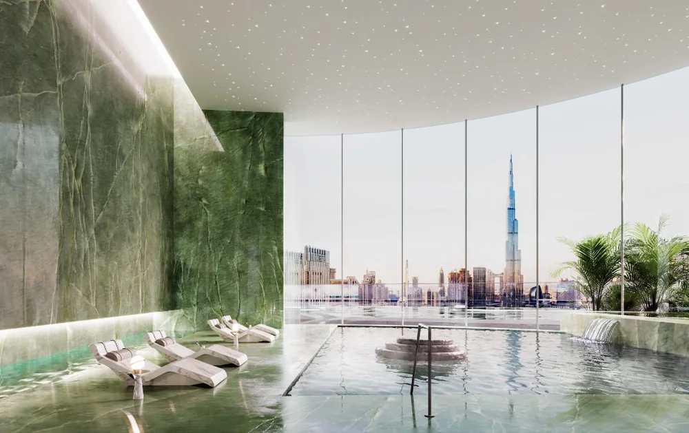Luxury indoor pool with green marble walls, lounge chairs, large windows with a cityscape view including the Burj Khalifa, and a small fountain feature.