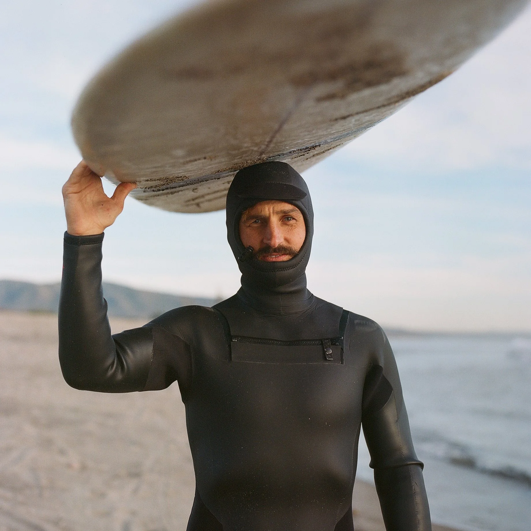 Man in a black wetsuit carrying a surfboard on his shoulder on the beach.