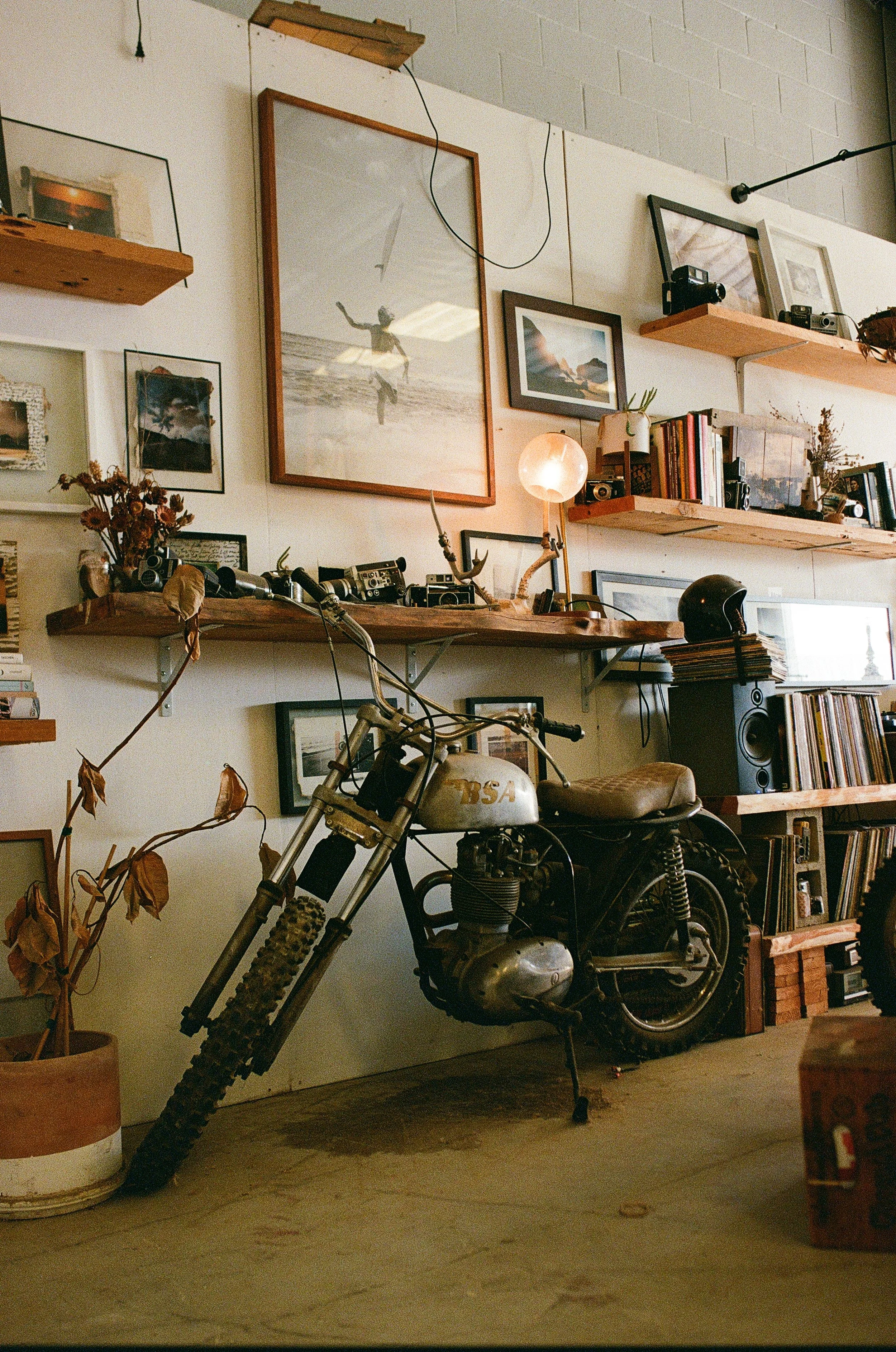 Vintage motorcycle in a cozy, eclectic room with wall-mounted shelves, framed photos and artwork, a lamp, stereo equipment, and dried flowers.