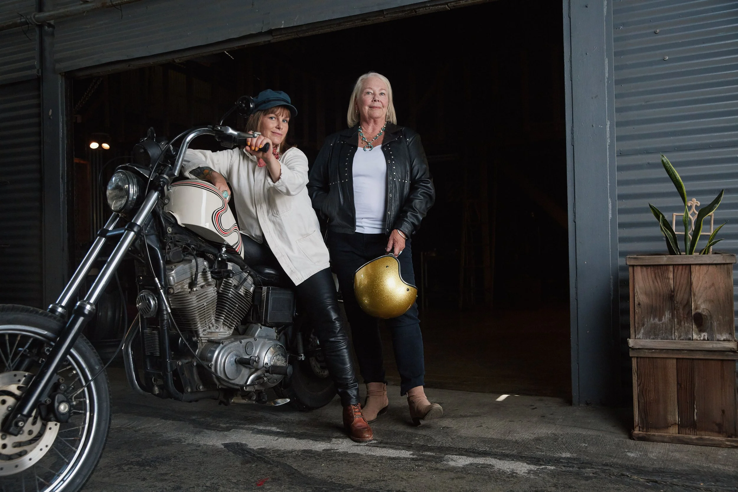 Two women standing near a motorcycle inside a garage or warehouse, one holding a helmet, the other resting her hand on the motorcycle, both dressed in casual leather and denim attire.