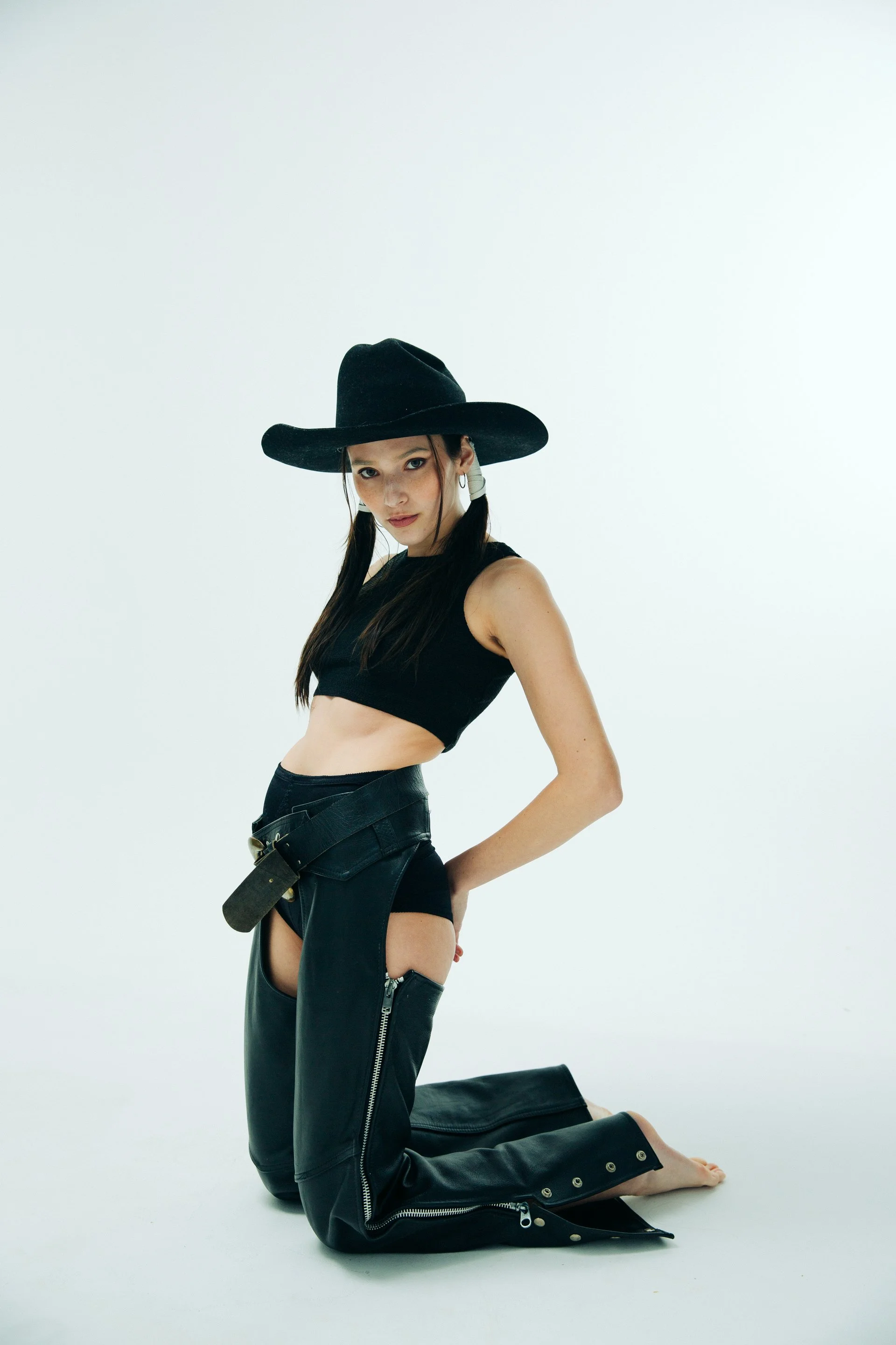 Young woman wearing a black wide-brimmed hat, black crop top, leather pants, and thigh-high leather boots, kneeling on the floor against a plain white background.