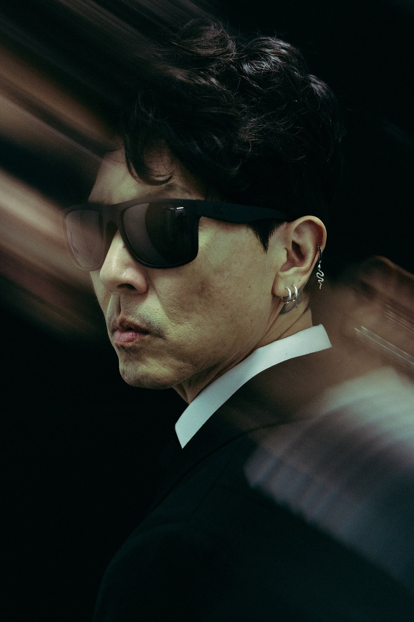 A man wearing black sunglasses, multiple earrings, and a black suit with a white shirt, looking sideways with a blurred background.