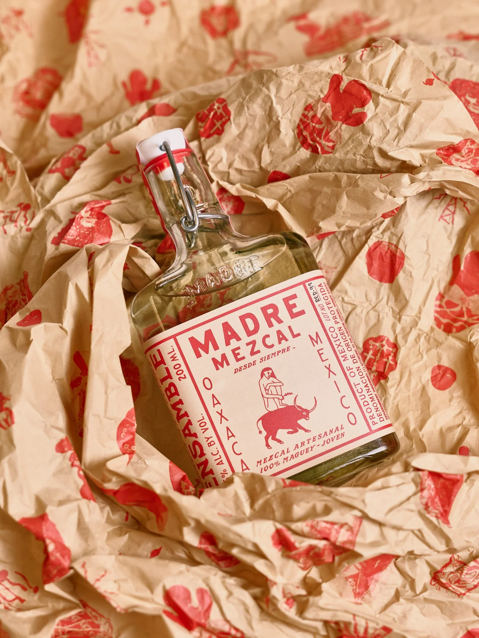 A small glass bottle of Madre Mezcal surrounded by crumpled brown paper with red printed logos.