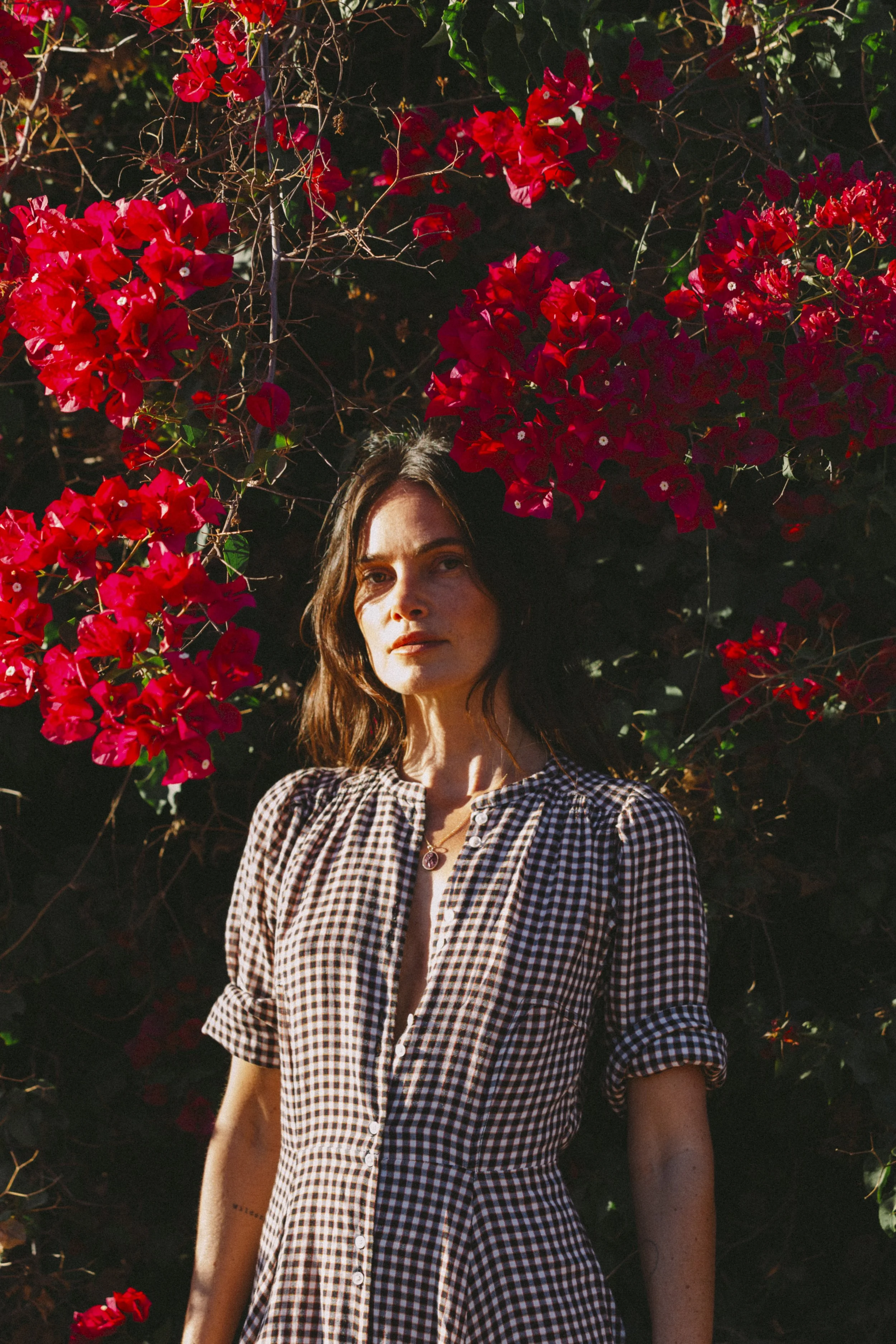 A woman with shoulder-length dark hair wearing a gingham dress standing in front of bright red bougainvillea flowers.