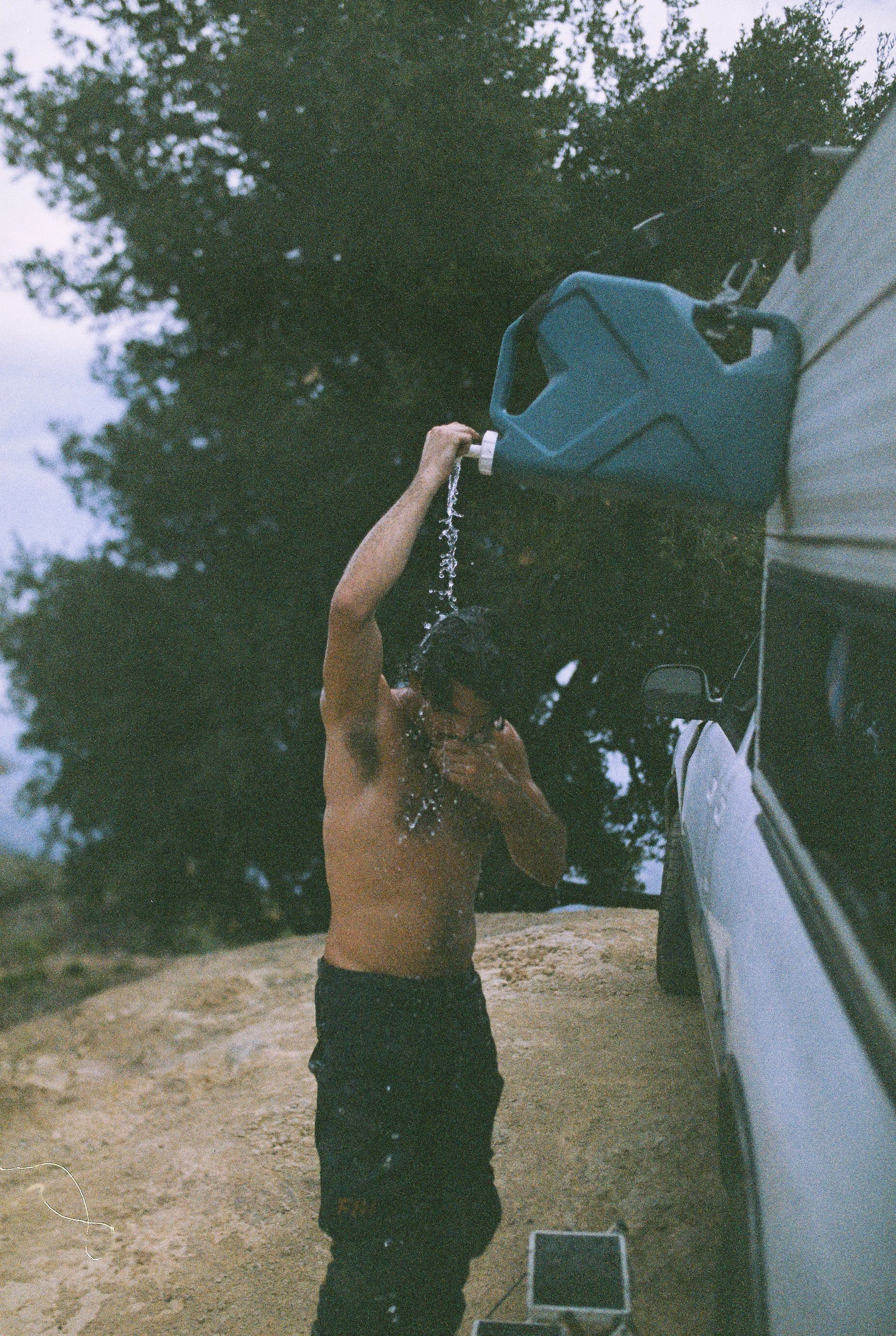 A man with dark hair, wearing black pants, is standing outdoors, pouring water from a blue plastic watering can onto his head, using a hook in his left hand to hold the can. The scene is set beside a silver vehicle on a dirt ground with trees in the 