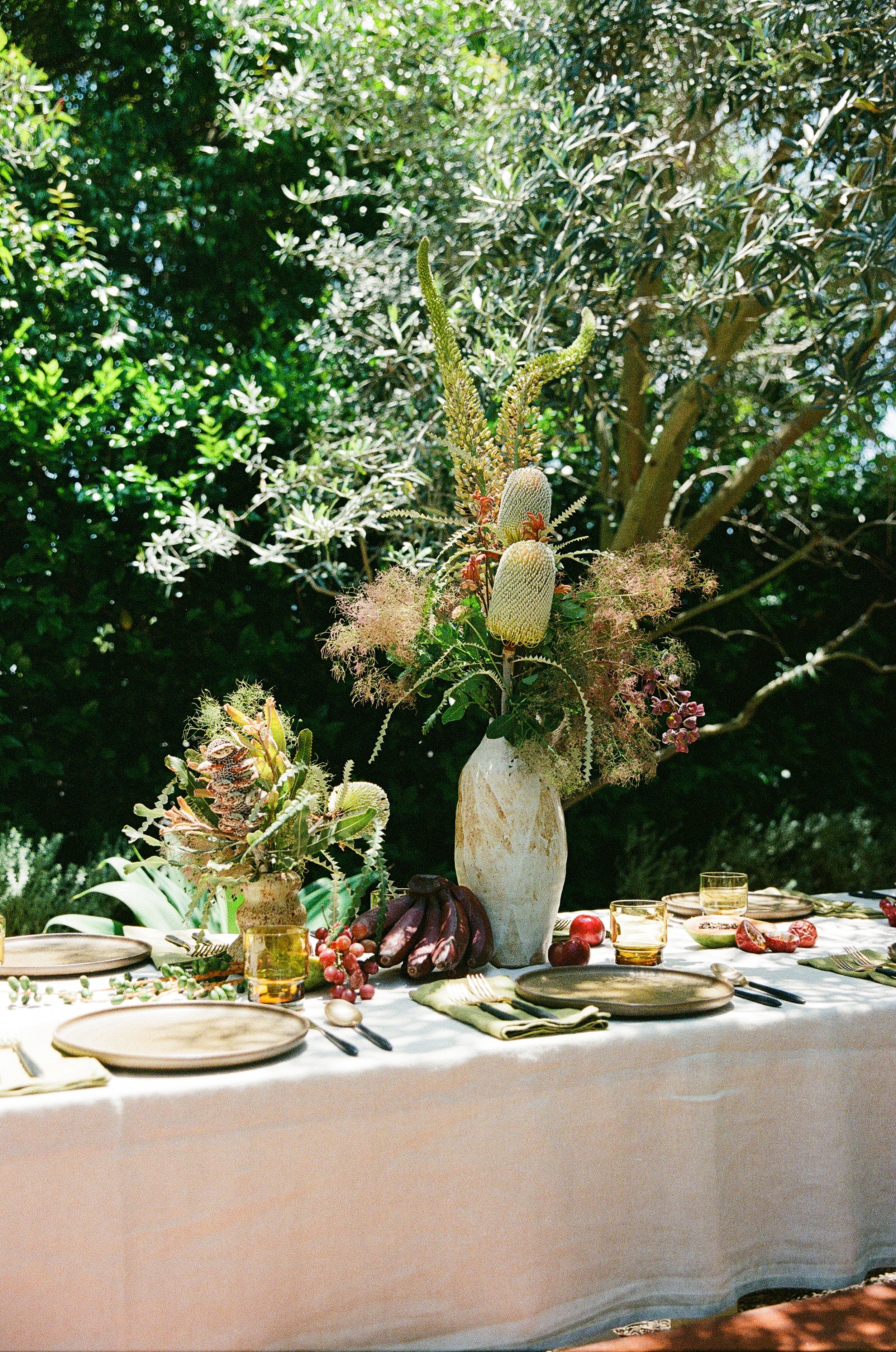 An outdoor dining table set with plates, glasses, utensils, and a large floral arrangement with various flowers and greenery, in a lush garden setting.