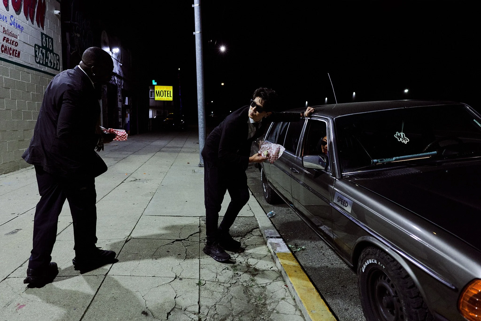 Two men in dark clothing and sunglasses sharing food from takeout containers outside at night, one leaning on a car parked along a cracked sidewalk, with a motel and neon signs in the background.